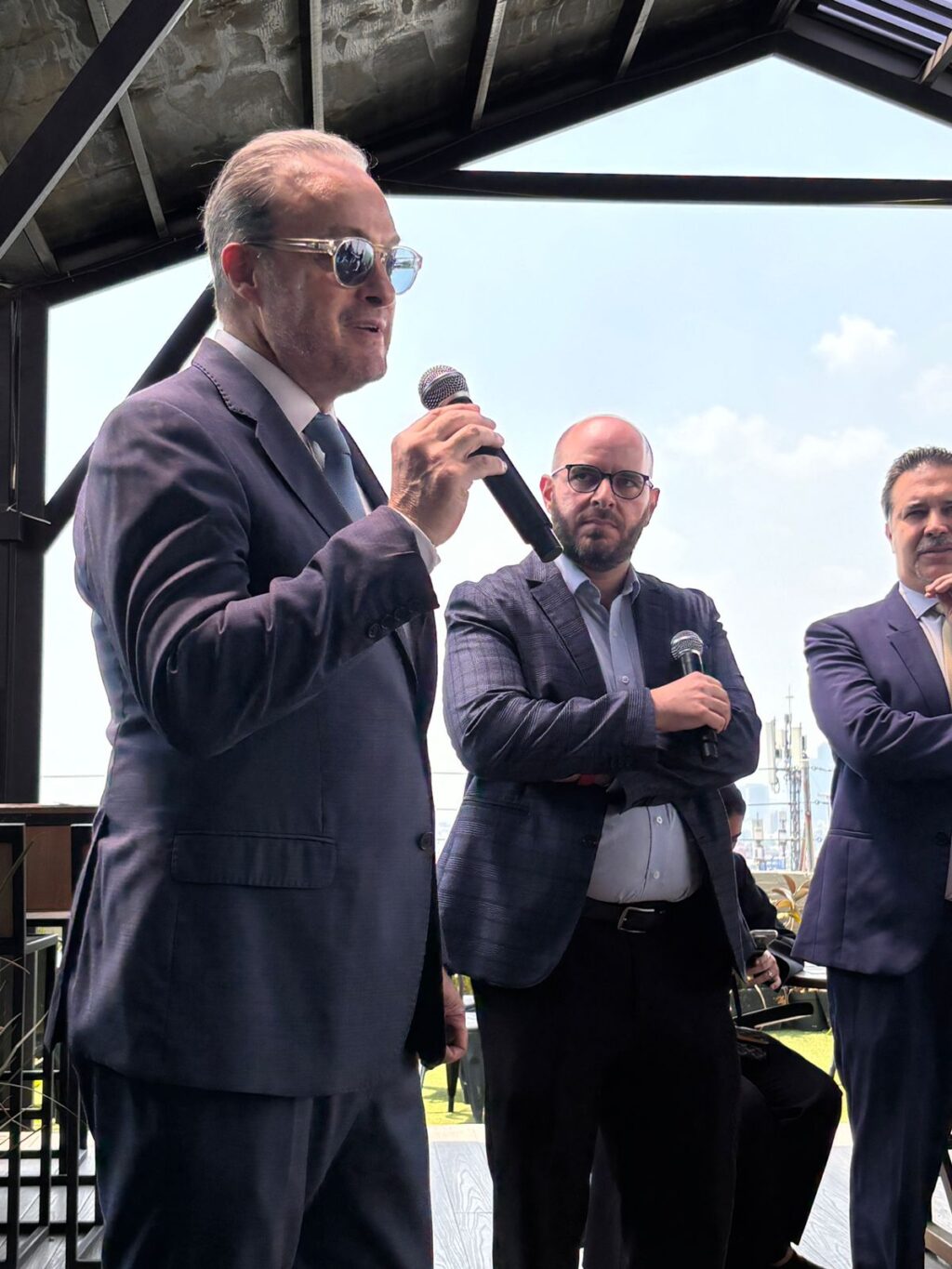 5cdb2a5e-a650-426c-8f94-6b6413aa05f0 - Petróleo y Energía Three men in suits on a stage; the foreground man speaks into a microphone while two colleagues listen, with a blue sky behind them.
