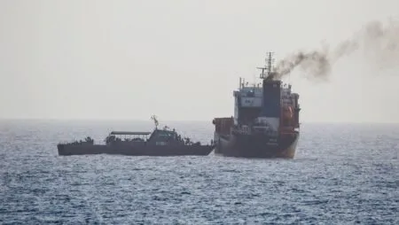 A tugboat beside a large container ship on open water, with a plume of smoke from the ship's stack.