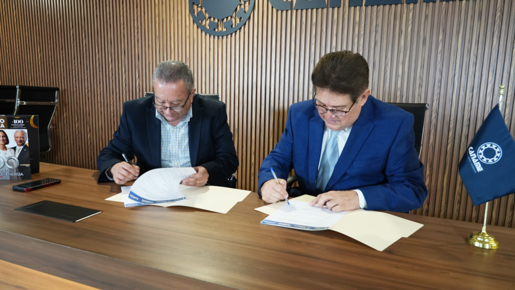 CANAME_01 - Petróleo y Energía Two men in suits sign documents at a wooden conference table during a formal meeting, with a blue flag nearby and a wood-paneled wall in the background.