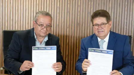 Two men in suits sit at a desk, each holding a printed document with logos, against a wood-paneled backdrop.