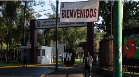 Entrance gate to an educational institute with a large 'BIENVENIDOS' banner overhead and a security booth nearby. A person walks toward the entrance on a tree-lined campus.