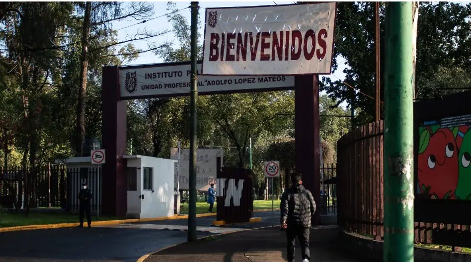 Entrance gate to an educational institute with a large 'BIENVENIDOS' banner overhead and a security booth nearby. A person walks toward the entrance on a tree-lined campus.