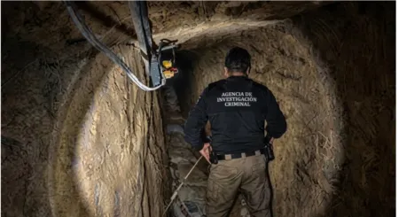Back view of a crime investigation agent wearing a black jacket with 'Agencia de Investigación Criminal' in a narrow underground tunnel, lit by a flashlight.