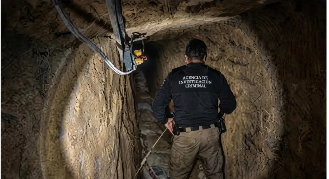 Back view of a crime investigation agent wearing a black jacket with 'Agencia de Investigación Criminal' in a narrow underground tunnel, lit by a flashlight.