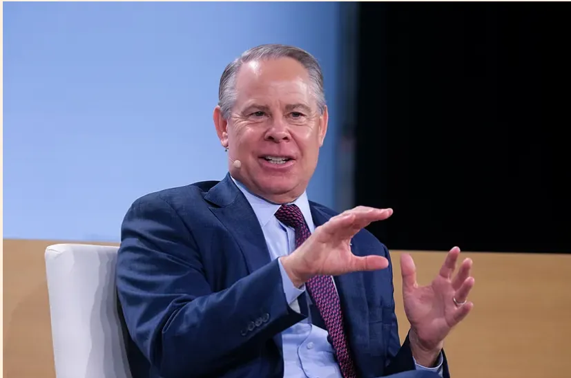 Man in a dark blue suit and red tie seated on stage, speaking with hands mid-gesture.