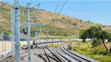 Railway tracks curve through a hilly landscape with a long white passenger train and workers inspecting the rails in the foreground. It shows overhead electric lines and a distant hillside.