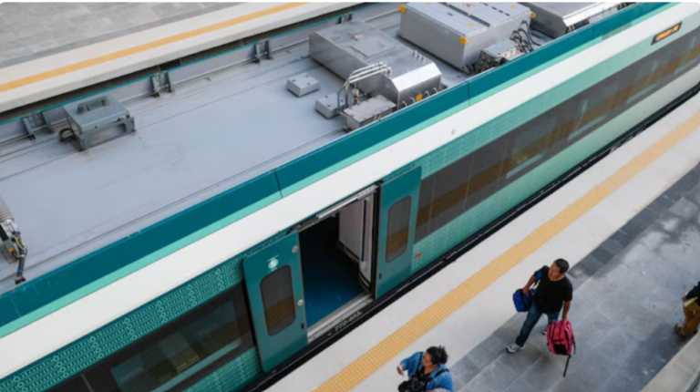Top-down view of a teal passenger train stopped at a platform, with two travelers carrying bags walking along the yellow tactile edge.