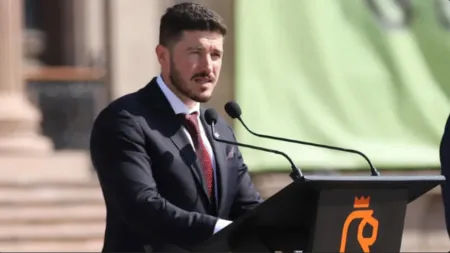 Man in a dark suit and red tie delivering a speech at a podium with two microphones outdoors.