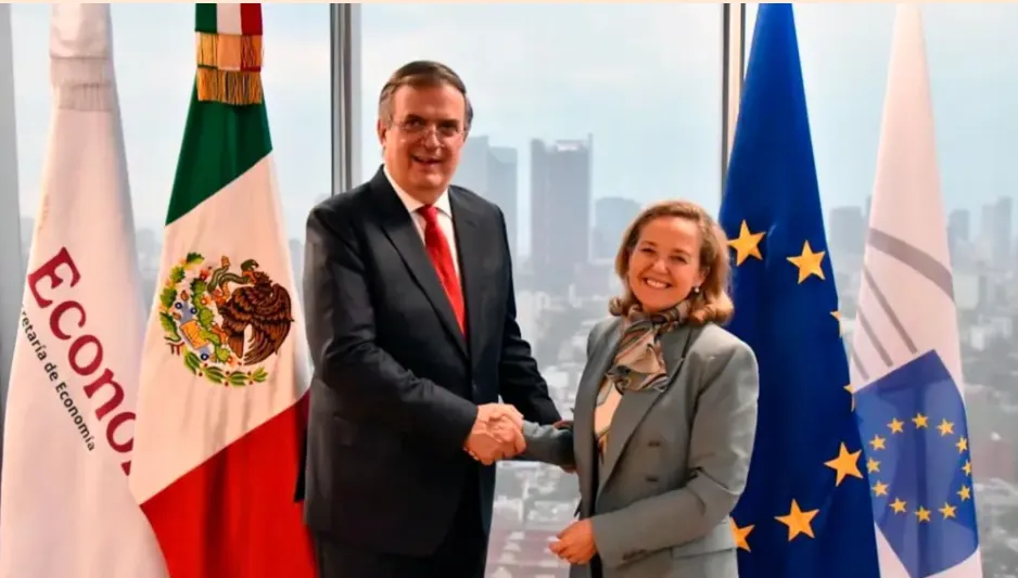 Two officials shake hands in front of flags, including Mexico and the European Union, smiling in a bright office setting.