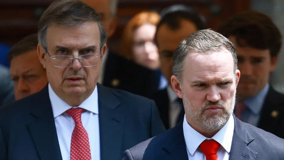 Two men in dark suits with red ties walk forward with stern expressions, a blurred crowd behind them.