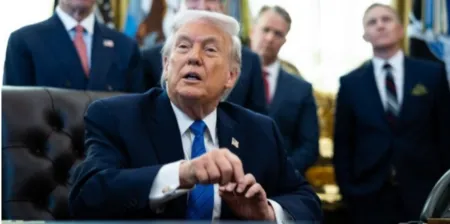 Man in a dark suit and blue tie sits at a desk in a formal office, aides in the background.