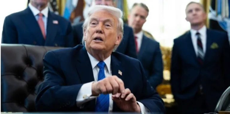 Man in a dark suit and blue tie sits at a desk in a formal office, aides in the background.