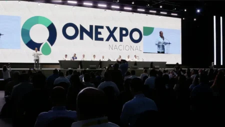 Speaker at a podium on a large stage with the ONEXPO NACIONAL logo backdrop and a long panel table behind, audience watching.