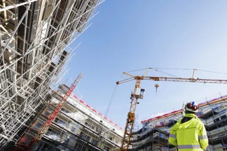 Construction site with metal scaffolding, a crane, and a worker in a high-visibility jacket against a blue sky.