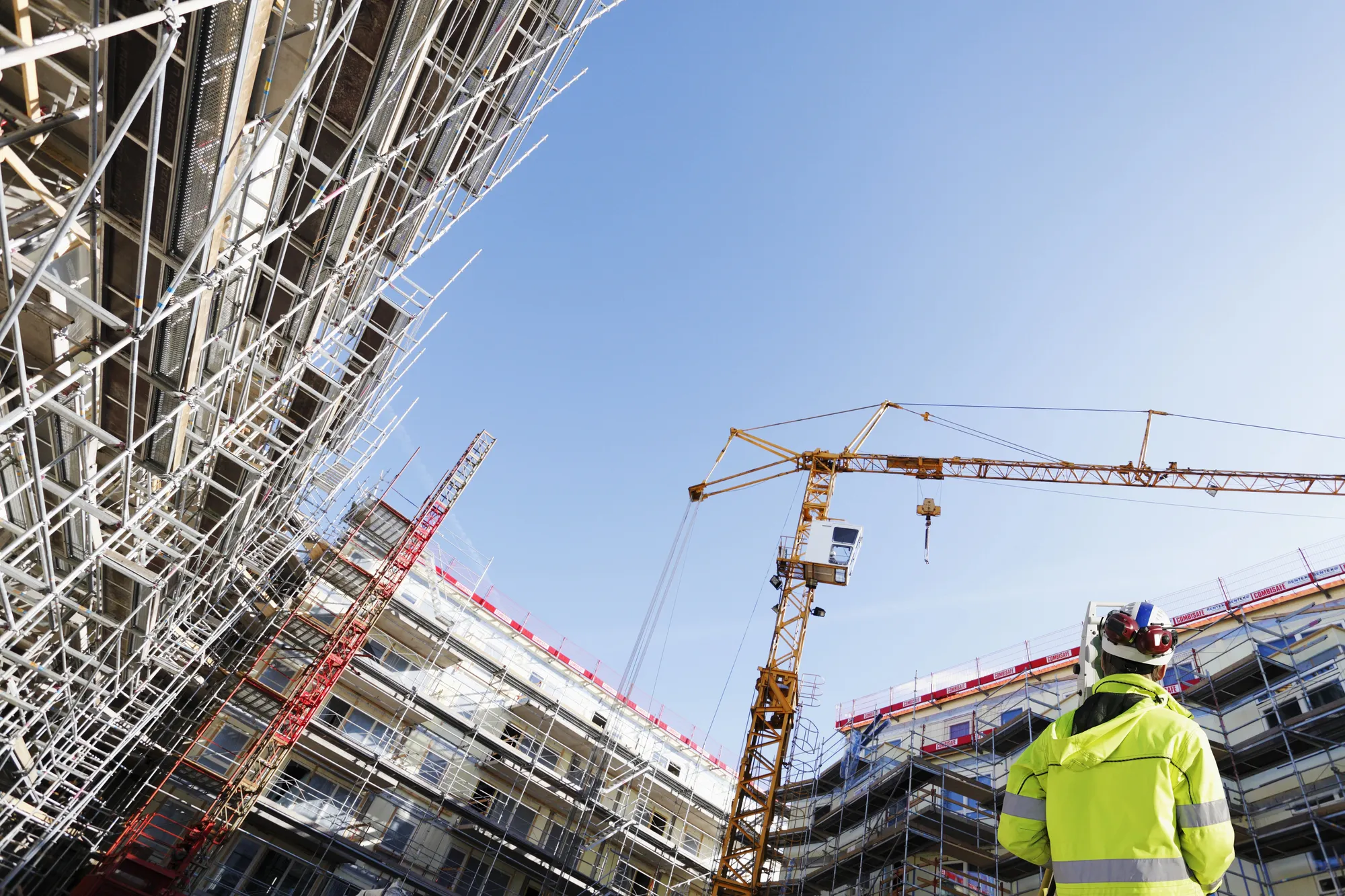 Construction site with metal scaffolding, a crane, and a worker in a high-visibility jacket against a blue sky.