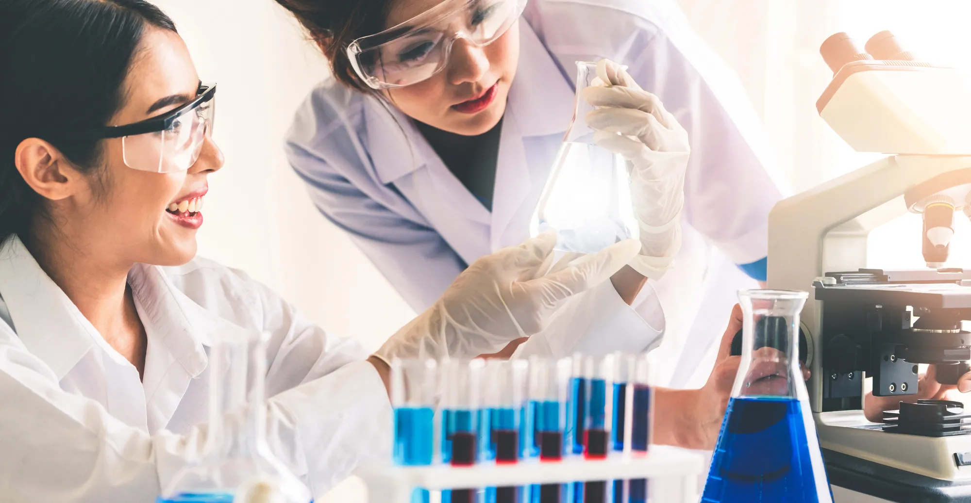 Two scientists in safety goggles and white coats hold an Erlenmeyer flask with blue liquid in a bright lab, with test tubes nearby.