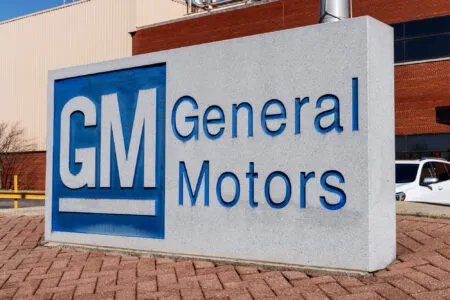 Concrete GM Motors sign with a blue General Motors logo and large lettering outdoors on brick and beige buildings.