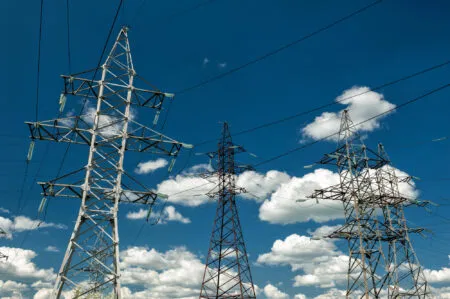 High-voltage transmission towers and wires against a bright blue sky with scattered clouds.