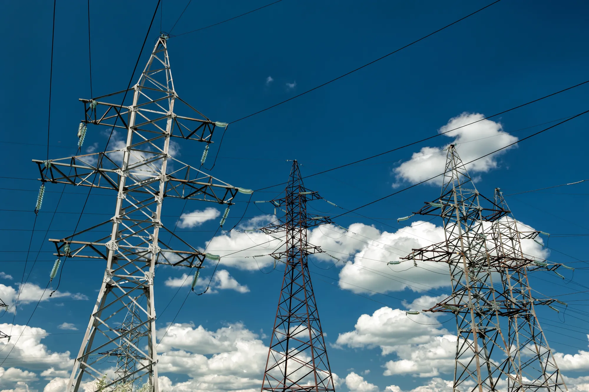 High-voltage transmission towers and wires against a bright blue sky with scattered clouds.