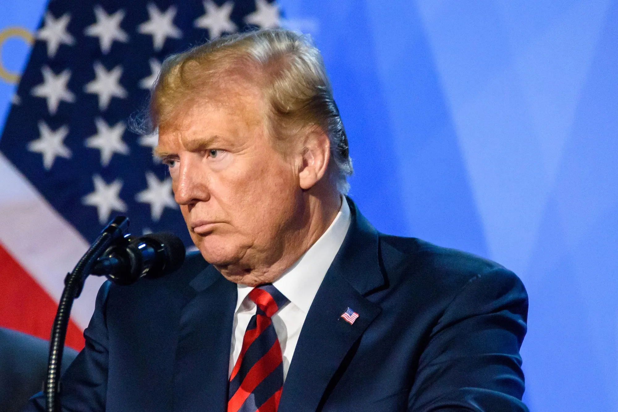 Politician at a podium in a dark suit and red striped tie, with an American flag backdrop behind him.