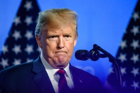 Close-up of a man in a dark suit and red tie speaking into a microphone against a blue backdrop with flag imagery behind him.