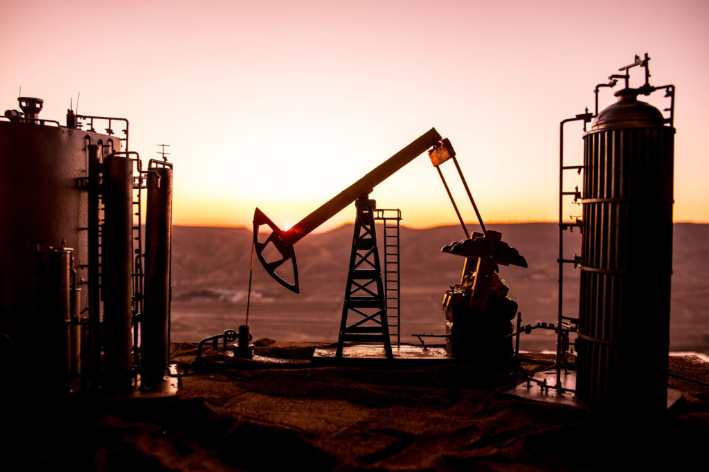 Silhouetted oil refinery equipment and pumpjacks against a warm sunset sky.