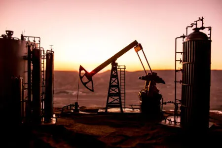 Silhouetted oil refinery equipment and pumpjacks against a warm sunset sky.