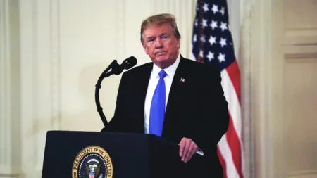 Man in a dark suit and blue tie delivering a speech at a podium with a U.S. presidential seal, an American flag in the background, and a microphone nearby.