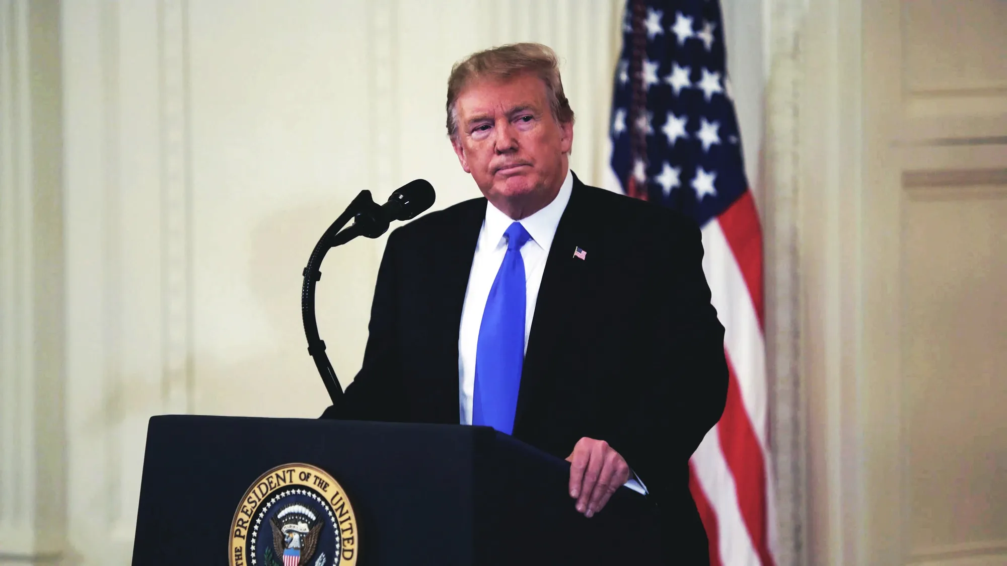 Man in a dark suit and blue tie delivering a speech at a podium with a U.S. presidential seal, an American flag in the background, and a microphone nearby.