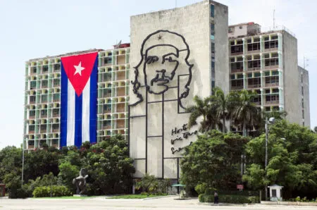 Tall building with a large Cuban flag banner on the left and a giant outline mural of Che Guevara on the central wall, with palm trees and a sculpture in the foreground.