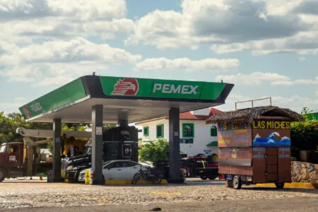 PEMEX gas station canopy with green branding under a blue sky, cars parked beneath it and a thatched-roof market stall nearby.
