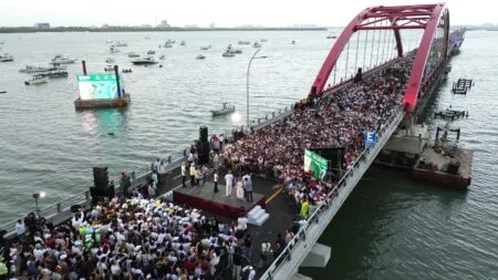 Crowd of people gathered on a pink arch pedestrian bridge during a public event, with a stage and speakers at the near end.