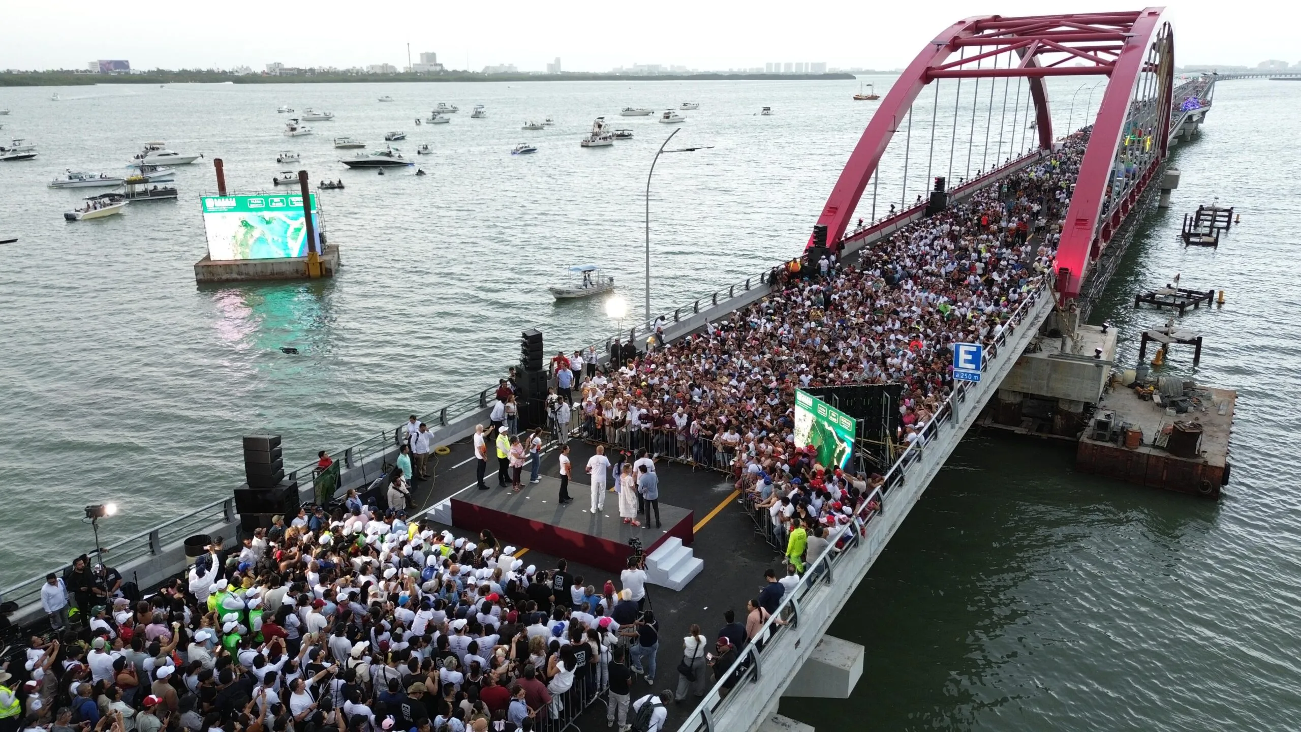 Crowd of people gathered on a pink arch pedestrian bridge during a public event, with a stage and speakers at the near end.