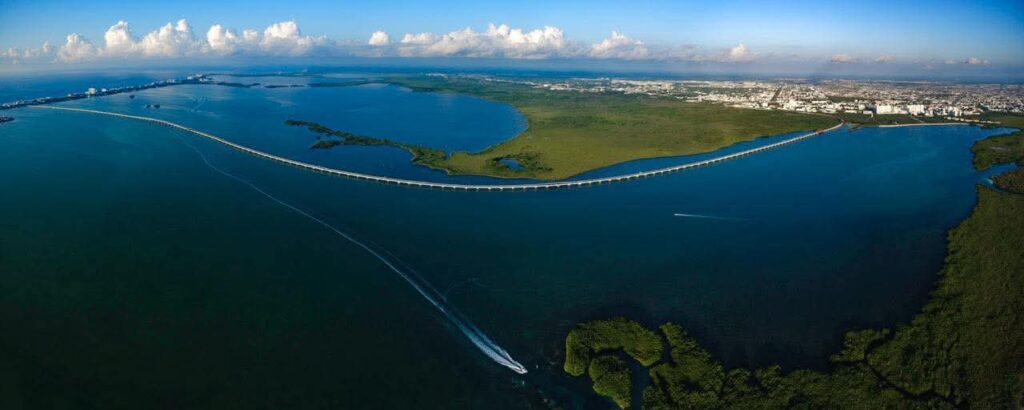 HHXm3wlboAAznAb - Petróleo y Energía Aerial view of a long curved bridge crossing a wide inlet, with green marshlands and a distant city horizon under blue skies.