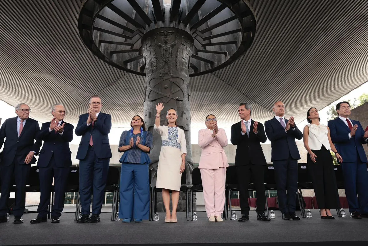 Line of formally dressed dignitaries standing on a stage, applauding, with a large circular metal sculpture overhead behind them.