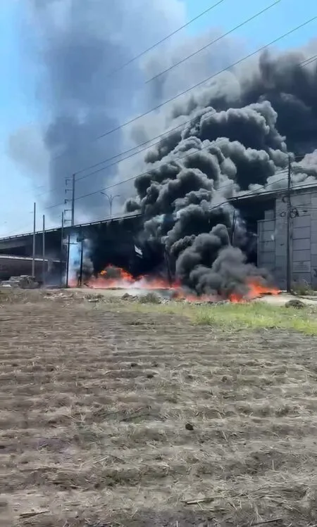 Industrial area with a large fire and thick black smoke billowing from a building/warehouse.