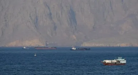 Calm blue sea with a small white boat in the foreground and several dark cargo ships near a rugged cliff coastline in the background.