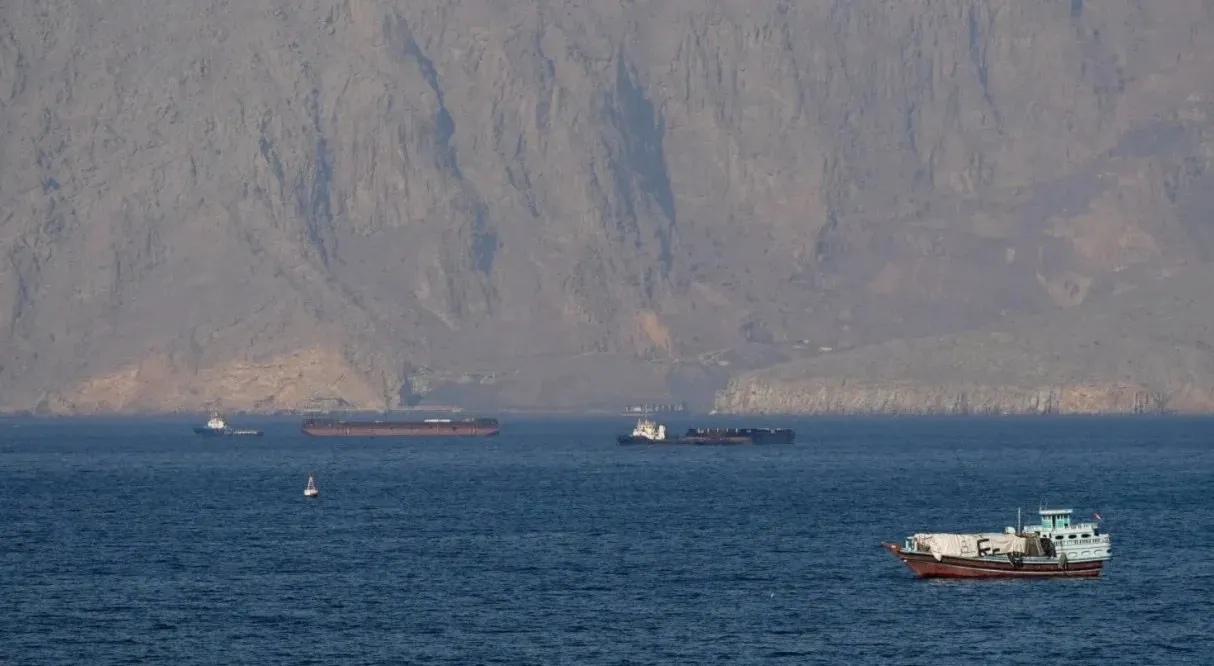 Calm blue sea with a small white boat in the foreground and several dark cargo ships near a rugged cliff coastline in the background.