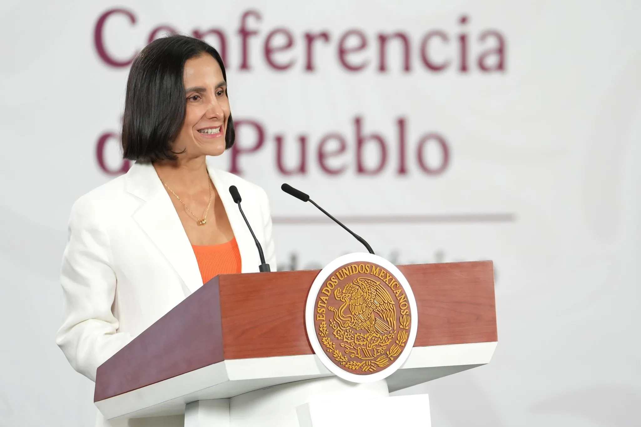 Woman in a white blazer speaks at a podium with the Mexican seal, microphones in front.