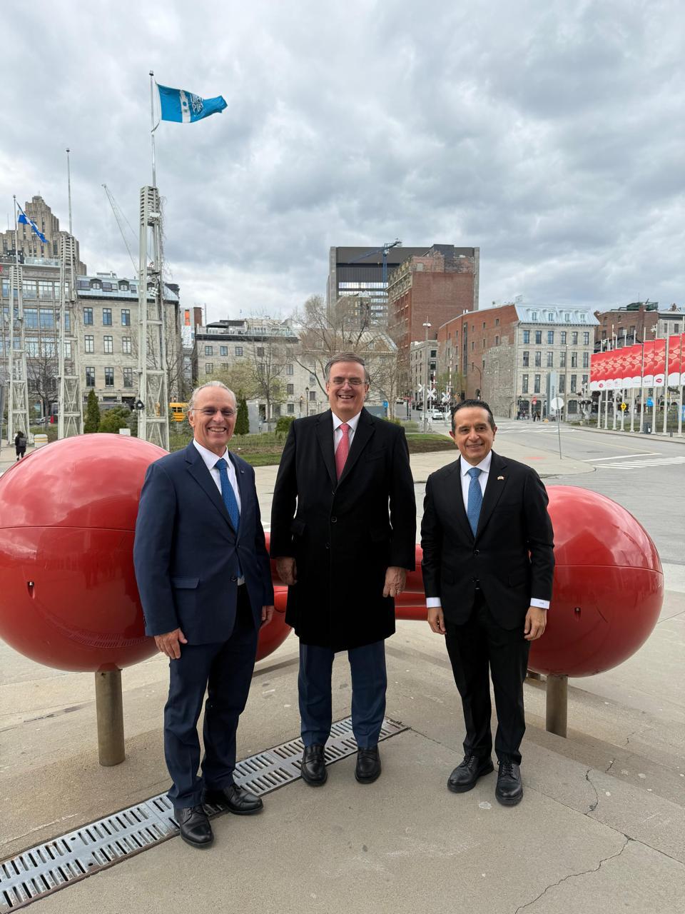 HHz-j0zW8AYryRA - Petróleo y Energía Three men in suits posing outdoors in front of a large red capsule sculpture in a city square.