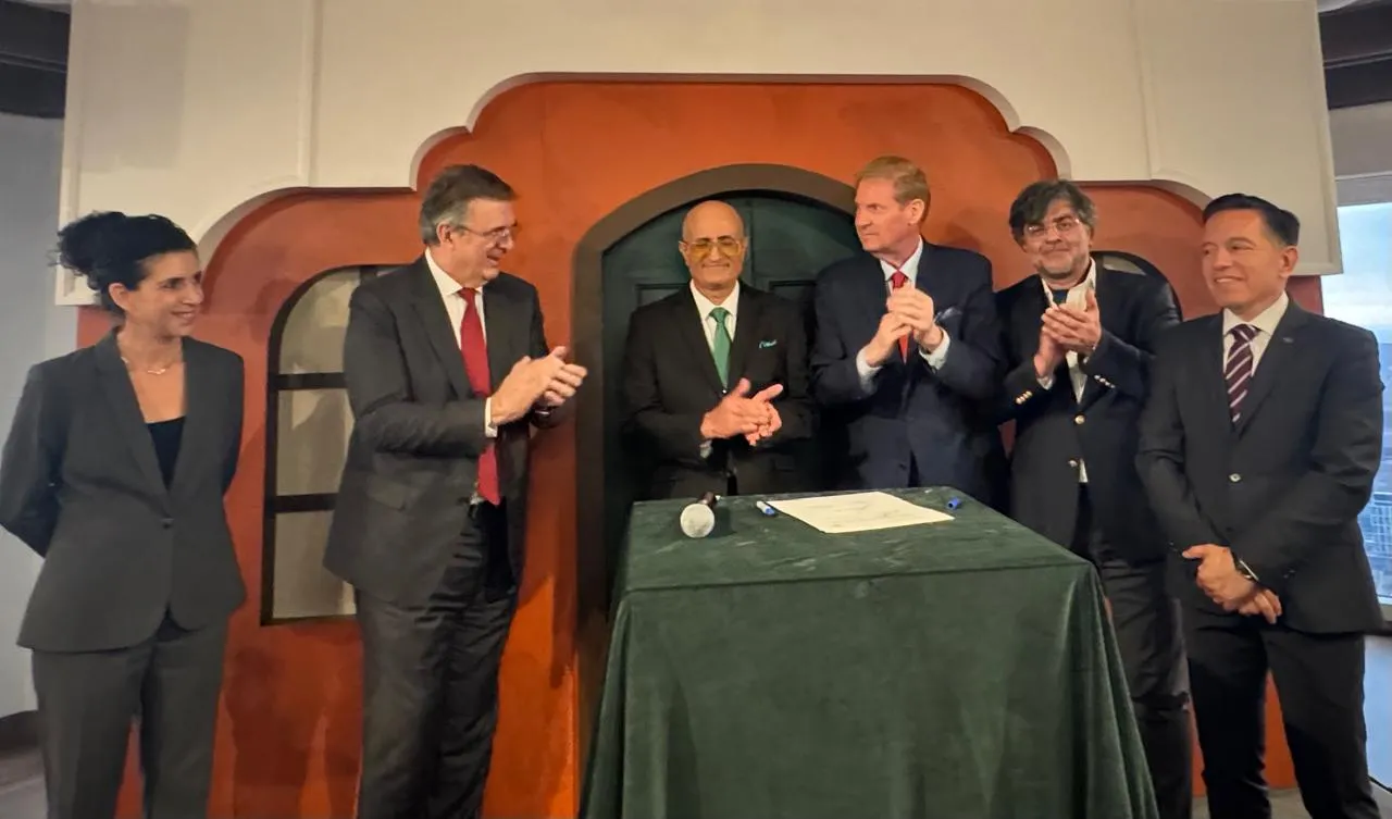 Six professionals in suits applaud around a table with a signed document during a formal ceremony.