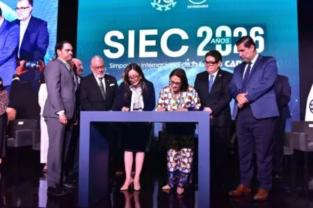 Six professionals in suits signing a document at a ceremony on a stage with a blue SIEC 2026 banner behind them.