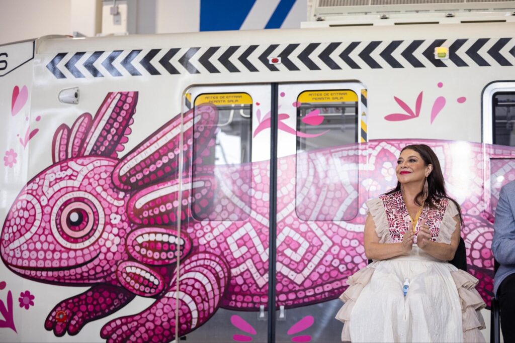 HID1A4CW8AAZTPM - Petróleo y Energía A smiling woman in a light-colored, ruffled dress stands beside a train car covered in bold pink graffiti with tentacle-like patterns on the side rough urban scene behind.