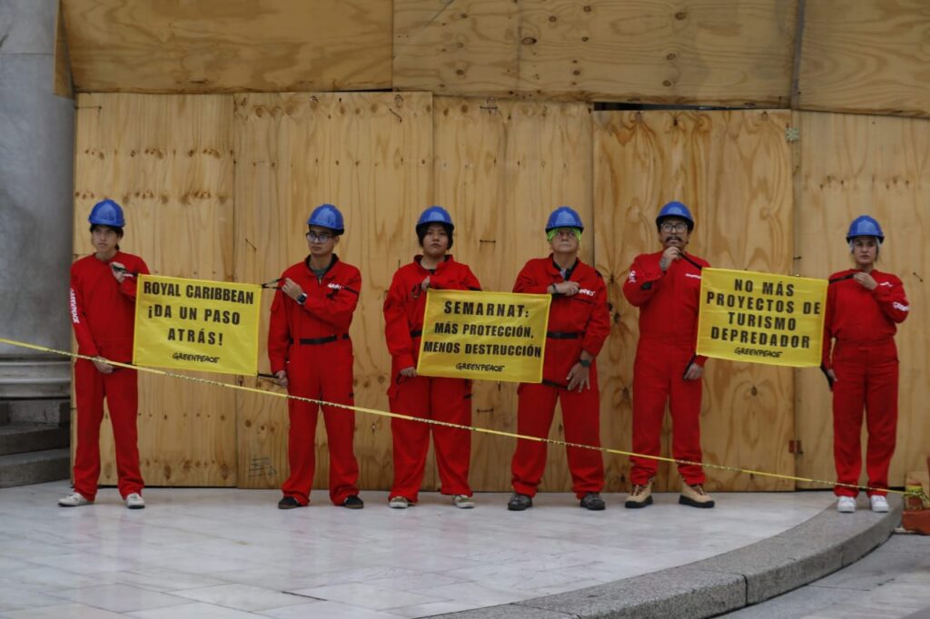 HII5-m_WwAAcSho - Petróleo y Energía Six Greenpeace protesters in red coveralls and blue helmets stand in front of plywood barriers, holding yellow signs urging action against Royal Caribbean and more environmental protection.",