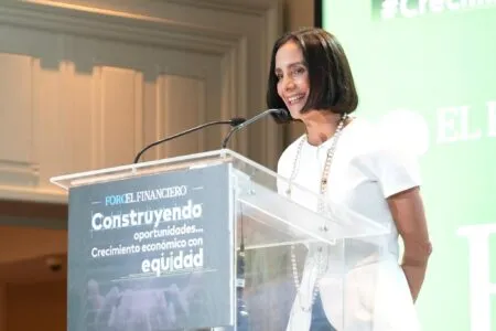 Professional woman in white speaking at a conference podium, smiling, with Foro El Financiero branding on the lectern and a green presentation backdrop behind her.