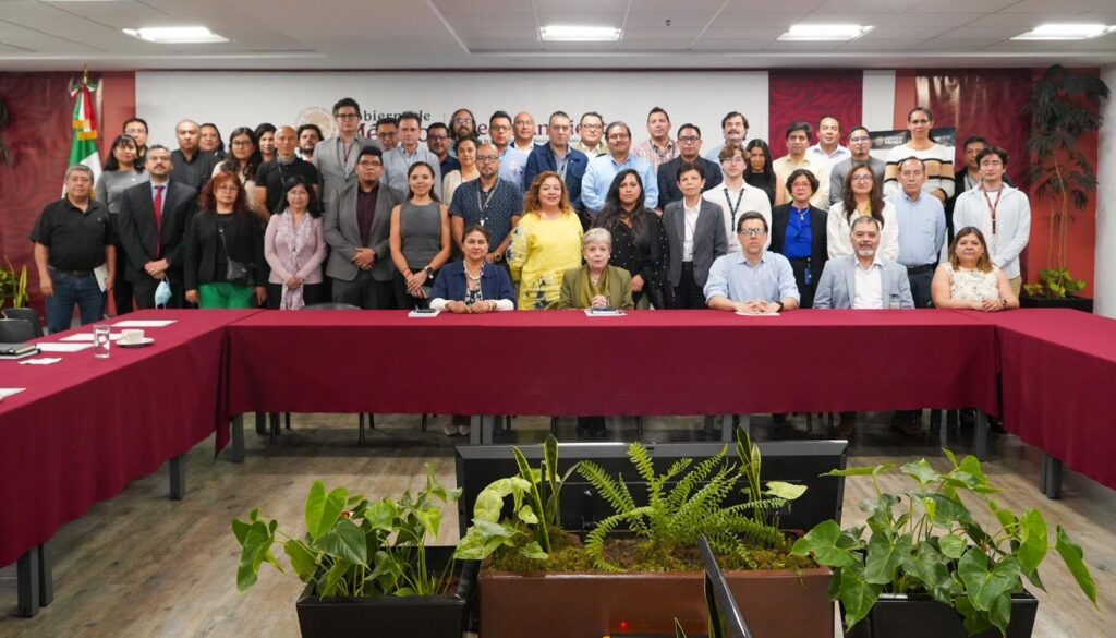 HJOKJv7XoAAczTv - Petróleo y Energía Group of professionals posing for a formal group photo behind a long red conference table in a meeting room, with a flag on the left and plants in the foreground.