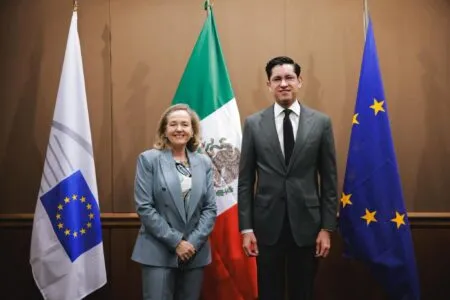 Two formally dressed officials stand side by side, smiling, with three flags behind them (a white flag with a blue emblem on the left, the Mexican flag in the center, and a blue flag with yellow stars on the right).