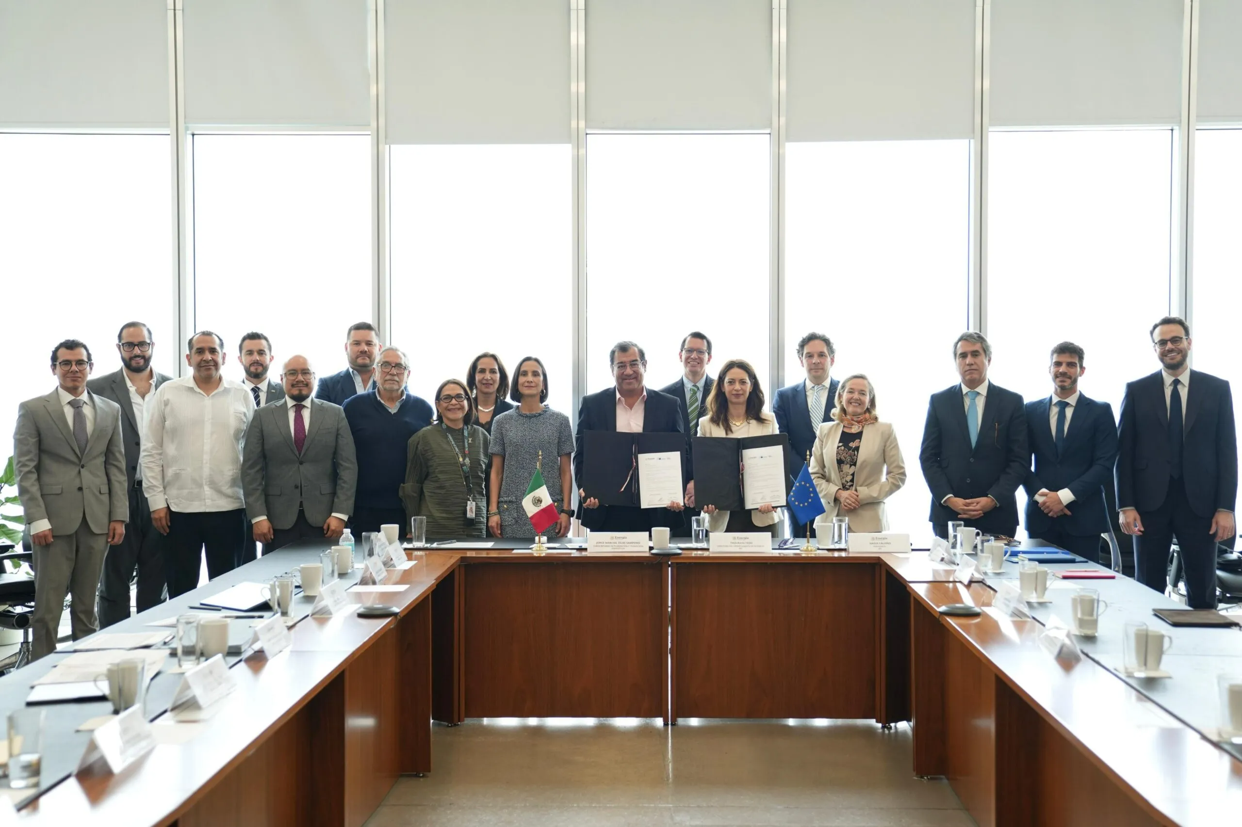Group of professionals in suits standing along a conference table with documents, flags (Mexico and EU) visible, posing for a photo in a bright office.