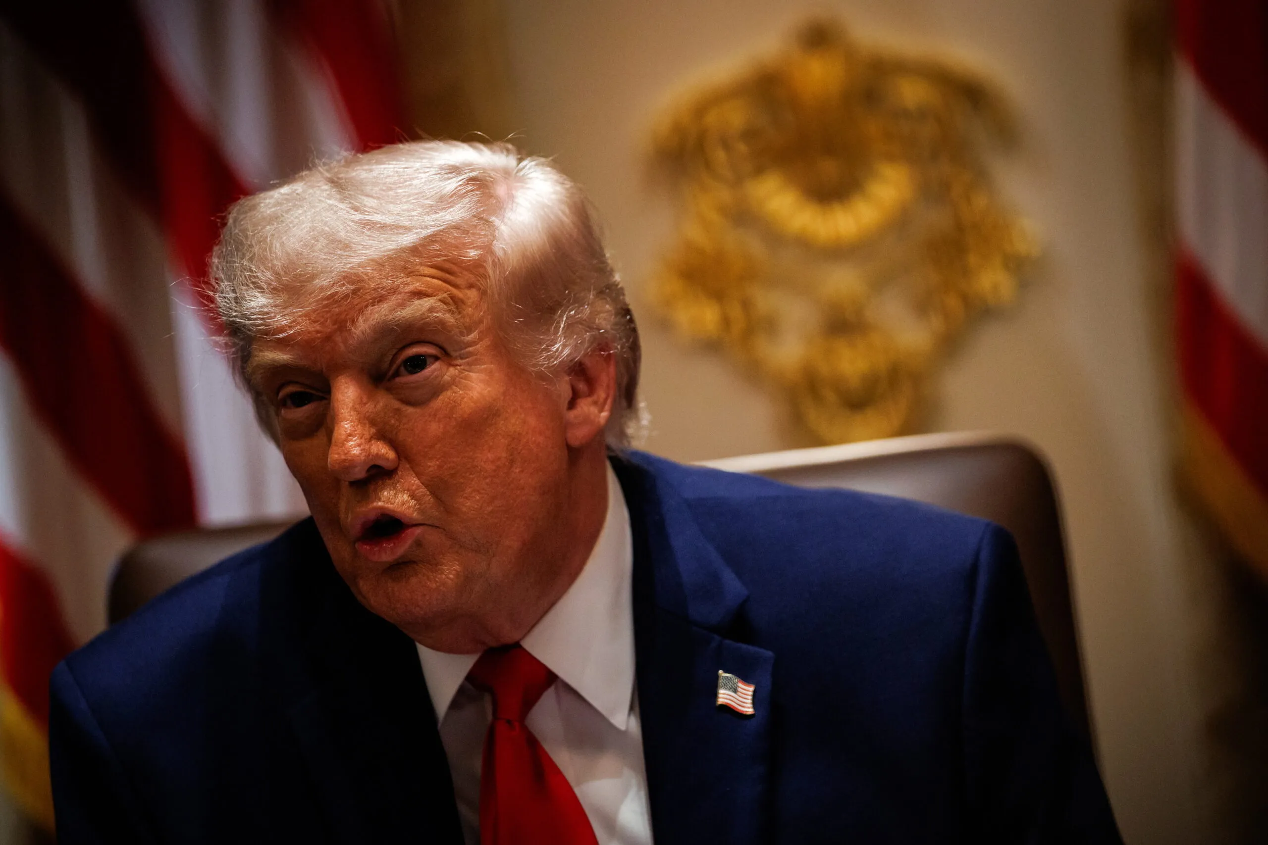 Close-up of a man in a blue suit and red tie, with a US flag pin, speaking with a blurred emblem in the background.