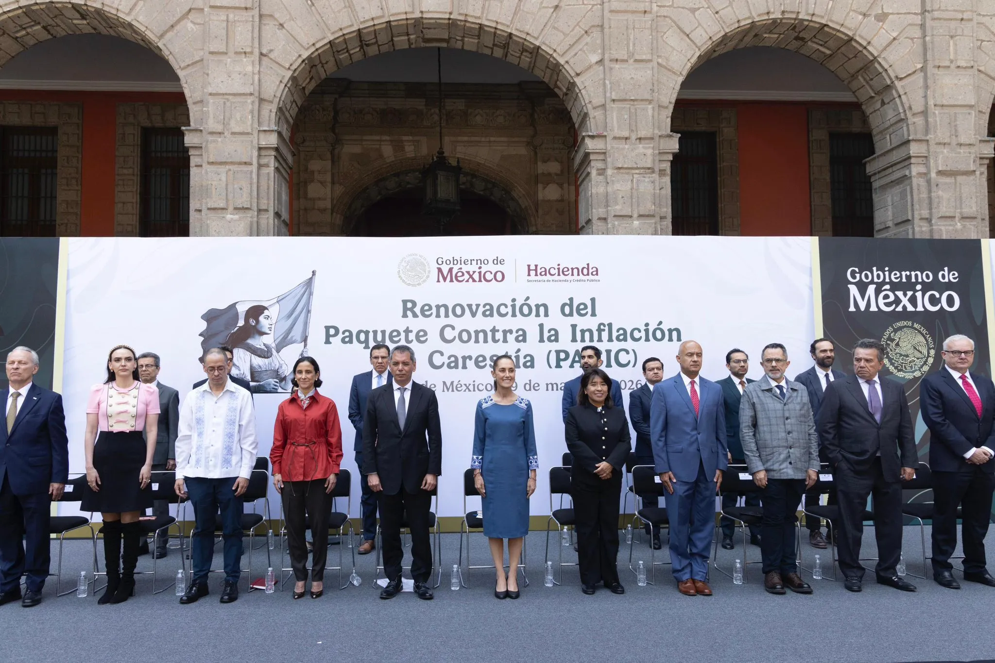 Group of formally dressed officials standing in front of a banner at a government event, with stone arches in the background